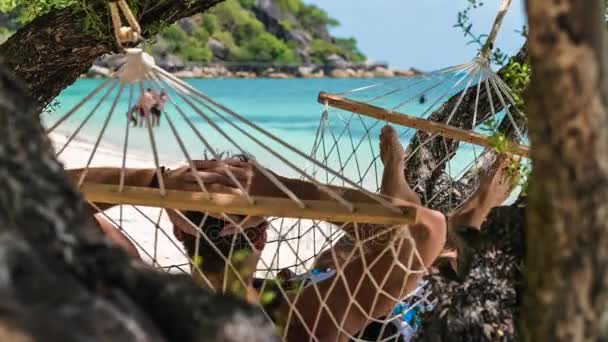 Güzel mavi okyanus önünde sahilde bir hamakta sallanan adam rahat. Onu güneşin Sihirbazı ağaçlarının gölgesi altında saklanıyor. Arka planda bazı turizm. — Stok video