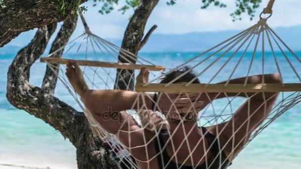 Man swingen ontspannen in een hangmat op het strand voor de blauwe oceaan. Verbergen van hem van de zon in de schaduw van de bomen van een wizard. — Stockvideo