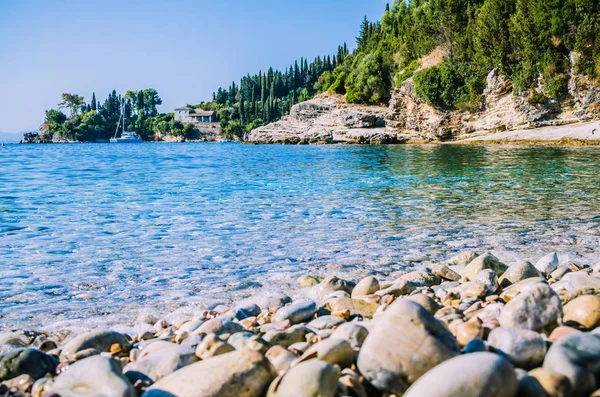 Spiaggia di ghiaia vicino Kalami con pini e cipressi e uno yacht all'ancora in una baia sullo sfondo. Isola di Corfù, Grecia — Foto Stock