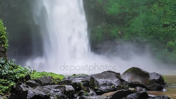 Close up de cachoeira Nungnung, Queda de água batendo superfície da água, algumas enormes rochas molhadas na frente do quadro. Lush green leafes motion in background, Bali, Indonésia — Vídeo de Stock