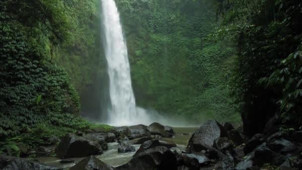 Удивительный водопад Нангнунг, медленно движущаяся падающая вода ударяется о поверхность воды, некоторые огромные скалы видны перед рамой. Пышные зеленые листочки движутся от ветра, Бали, Индонезия — стоковое видео