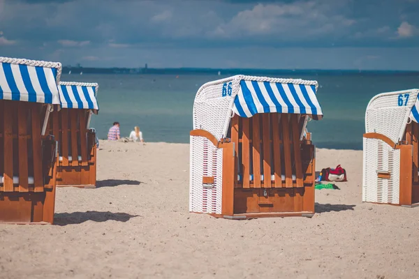 Cadeiras com telhados listrados coloridos na praia de areia em Travemunde. Um casal desfocado sentado na praia ao fundo. Lubeck, Alemanha — Fotografia de Stock