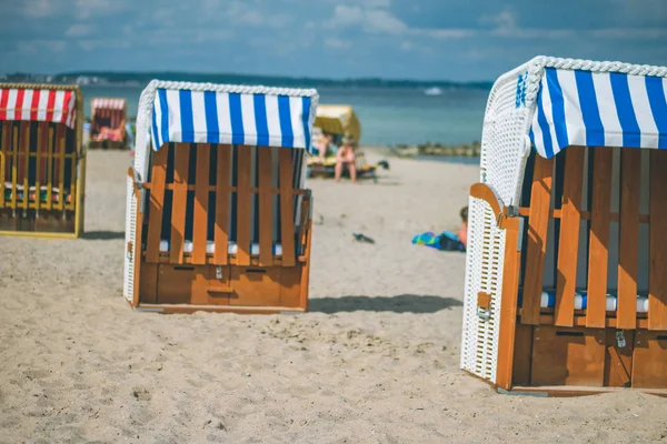 Cadeiras com telhados listrados coloridos na praia de areia em Travemunde. Um casal desfocado sentado na praia ao fundo. Lubeck, Alemanha — Fotografia de Stock