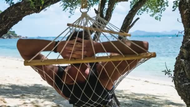 Homem balançando relaxado em uma rede na praia em frente ao belo oceano azul e outra ilha no fundo. Escondendo-se do sol à sombra de uma árvore . — Vídeo de Stock