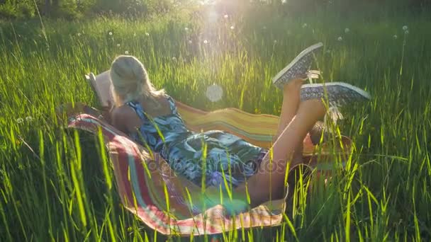 Une jeune fille blonde allongée sur une couverture dans une herbe classée et lue livre. Coucher de soleil rétroéclairé dans le cadre. Vue arrière. Mouvement manuel — Video