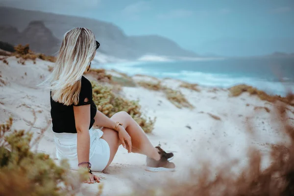 Mulher sentada em uma duna de areia branca com vegetação estéril admirando a paisagem costeira e as ondas oceânicas atlânticas. São Vicente Cabo Verde — Fotografia de Stock