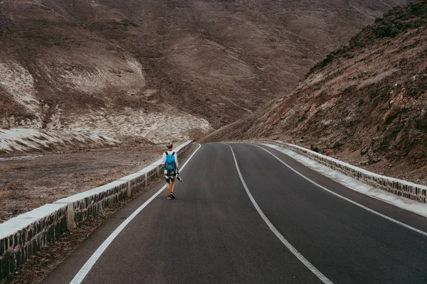 Путешествующая женщина с рюкзаком ходит по пустой дороге среди скалистых вулканических ландшафтов Огромные вулканические горы поднимаются с обеих сторон. Сан-Висенте-Кабо-Верде — стоковое фото