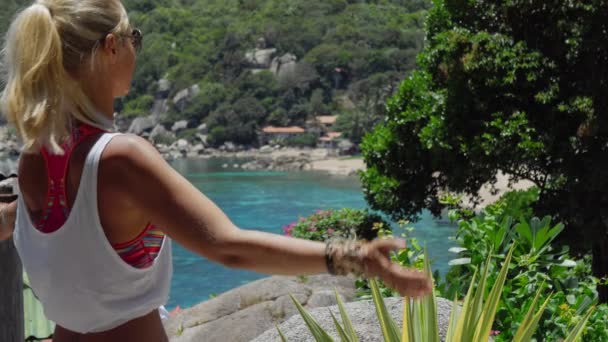 Mujer deportiva bronceada meditando frente a la hermosa bahía de Tanote en un día soleado. Ligera brisa del océano brisa del océano tamizada a través de su cabello rubio. Koh Tao, Tailandia — Vídeos de Stock