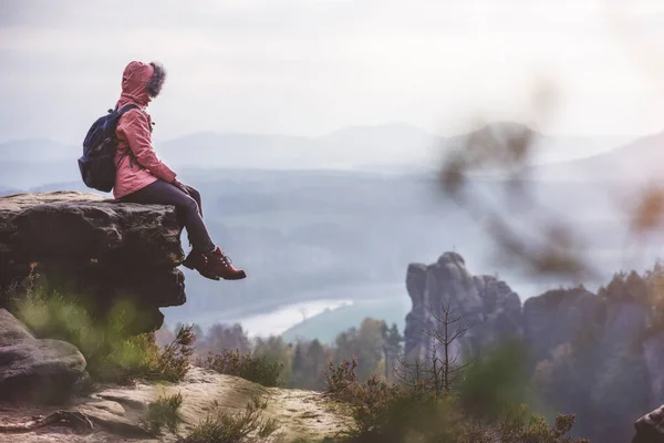 Jovem mulher em roupas ao ar livre com mochila sentada na borda dos penhascos desfrutando de vista do cume da montanha, floresta e rio no vale. Conceito de estilo de vida — Fotografia de Stock