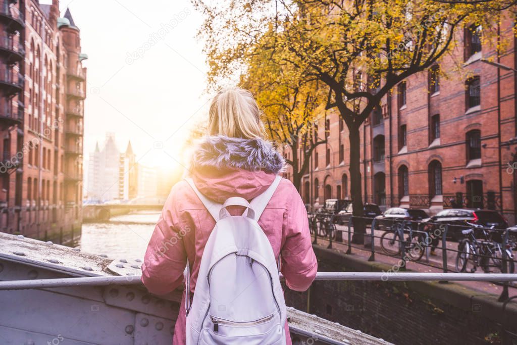 Rear view of tourist adult blond woman with backpack enjoying beautiful sunset scene on the Speicherstadt historic warehouse district in Hamburg, Germany, Europe