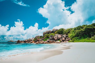 Seyşeller 'deki La Digue adasındaki Grand Anse egzotik, el değmemiş plajı. Beyaz kumlu sahil, mavi okyanus lagünü, dalgalar, beyaz bulutlar, arka planda granit kayalar.