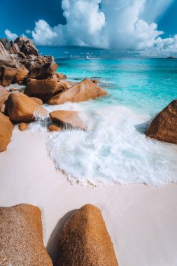 Grand Anse sahilindeki kayaların arasında köpük dalgası, La Digue, Seyşeller. Beyaz bulutlar, berrak okyanus ve yalnız lüks tekne.