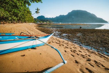 Bangka banca teknesi kumlu kumsalda altın günbatımı ışığıyla aydınlatılıyor. Arka planda El Nido köyü. Filipinler