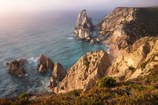 プライア・ダ・ウルサ・ビーチの夕日。大西洋の海のスタックとロッキー崖の尾根は、夜暖かい光で照らされています。シントラ地方,ポルトガルでの休暇 — ストック写真