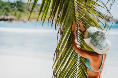 Hindistan cevizi palmiyesinin yanında güneş şapkası takan bir kadın tropik bir Anse Cocos kumsalında yaprak çırpıyor. La Digue, Seyşeller