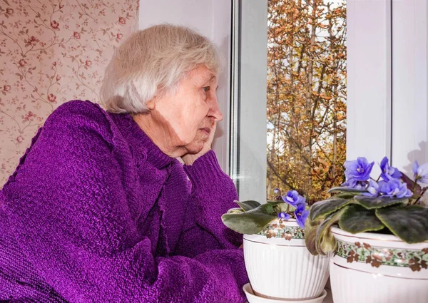 Vieille femme solitaire assise près de la fenêtre dans sa maison. — Photo
