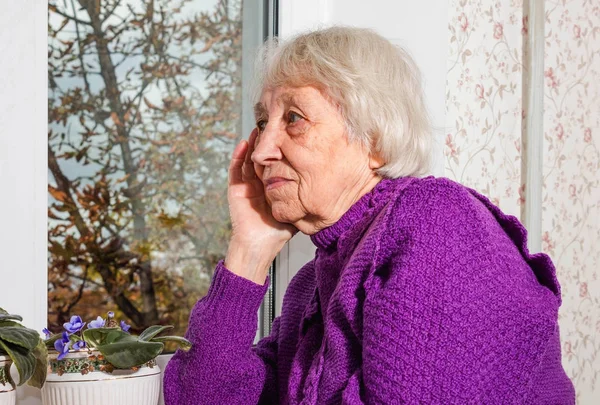 Old lonely woman sitting near the window in his house. — Stock Photo, Image