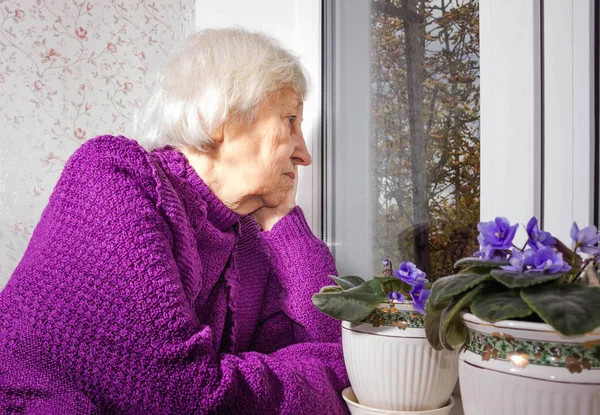 Old lonely woman sitting near the window in his house. — Stock Photo, Image