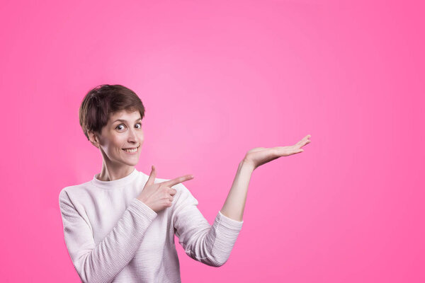 Excited girl pointing showing at pink background with copy space. Young beautiful happy smiling and laughing woman