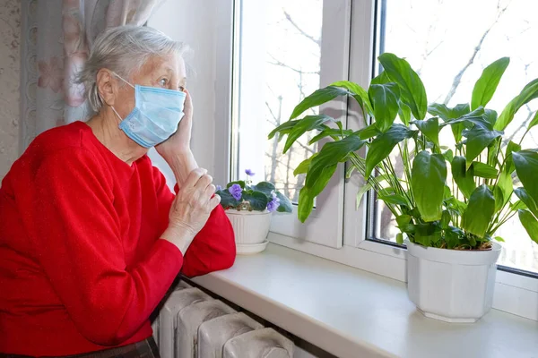 The Covid-19, health, safety and pandemic concept - senior old lonely woman wearing protective medical mask sitting near the window in his house for protection from virus