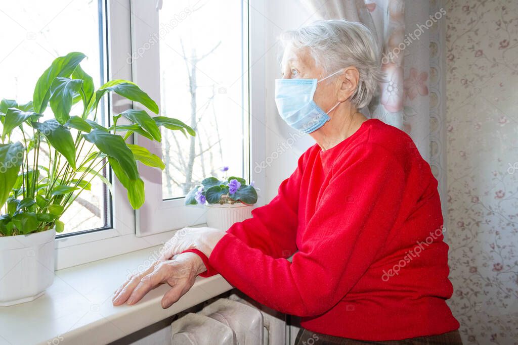 The Covid-19, health, safety and pandemic concept - senior old lonely woman wearing protective medical mask sitting near the window in his house for protection from virus