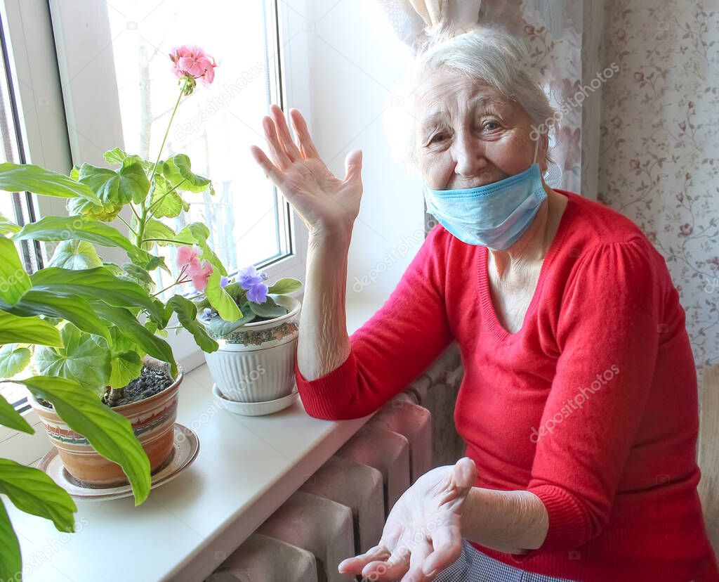 So, here we are. The Covid-19, health, safety and pandemic concept - senior old lonely woman wearing protective medical mask sitting near the window at home for protection from virus