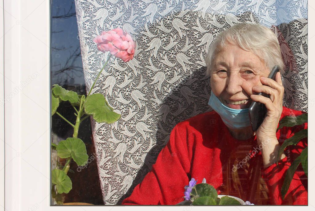 The Covid-19, health, safety and pandemic concept - senior old lonely woman wearing protective medical mask sitting near the window at home for protection from virus with mobile phone