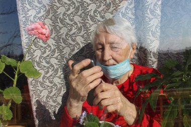 Covid-19, sağlık, güvenlik ve salgın konsepti - koruyucu maske takan yaşlı, yalnız kadın cep telefonlu virüsten korunmak için evdeki pencerenin yanında oturuyor.