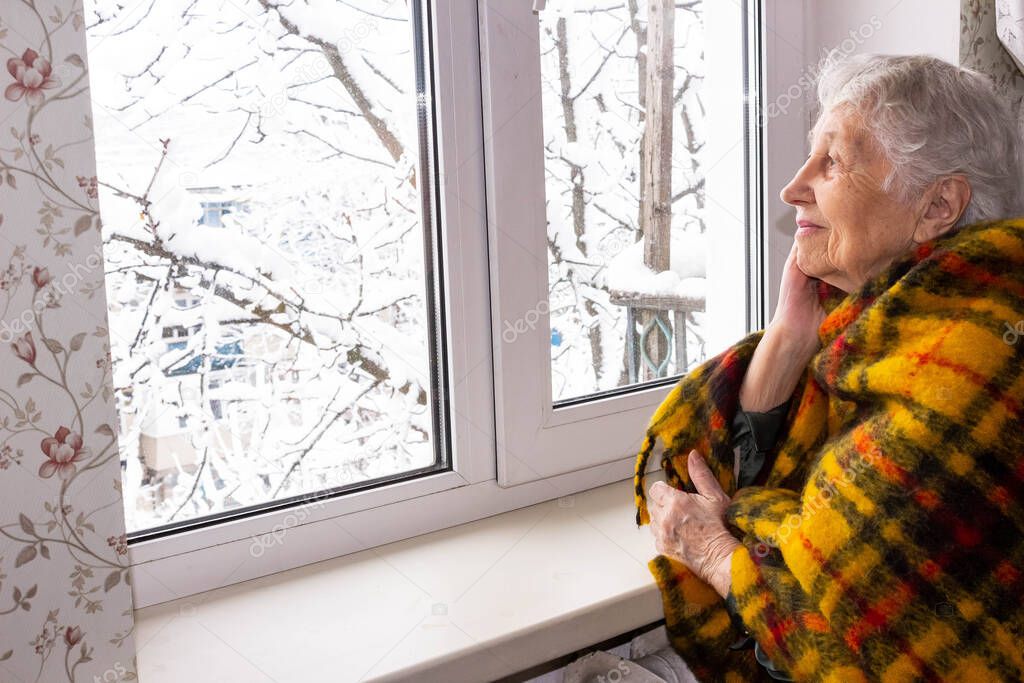 Old lonely woman sitting near the window in his house and looking and dreaming at winter