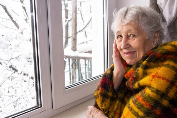 Old lonely woman sitting near the window in his house and looking at camera and smiling at winter