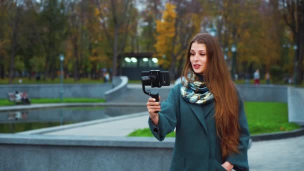 Девушка-видеоблоггер держит стабикам со смартфоном и в прямом эфире из парка — стоковое видео
