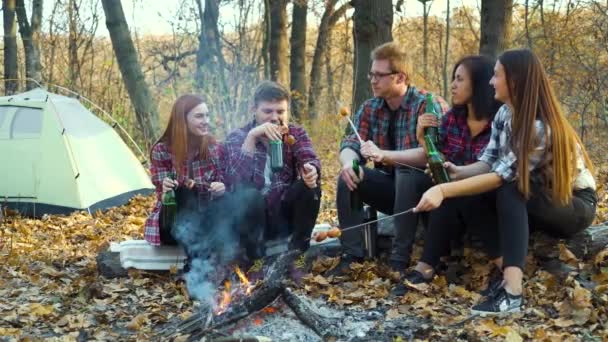 Felices turistas bebiendo cerveza y comiendo salchichas junto a la fogata en el bosque de otoño — Vídeos de Stock
