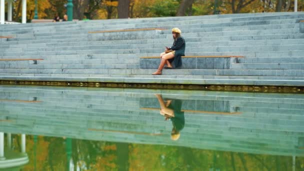 Самотня дівчина смс-повідомлення, що сидить на сходах біля ставка — стокове відео