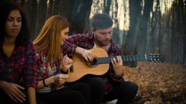 Genç turist, sonbahar ormanlarında kamp ateşi eşliğinde gitar akort ediyor.