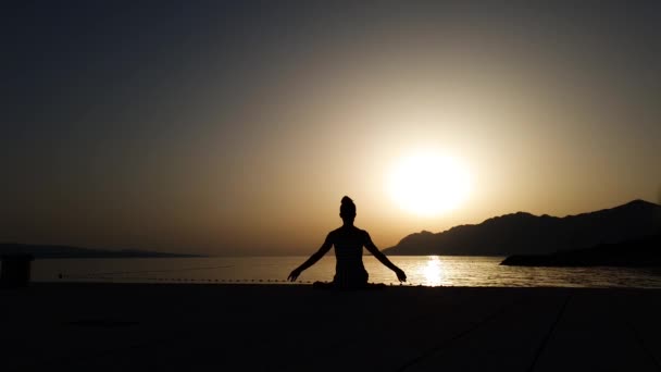 Vreedzaam meisje zit in yoga poseren bij zonsondergang door de zee — Stockvideo