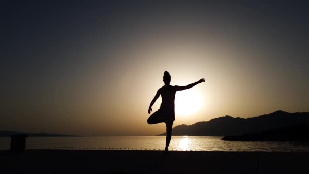 Yogacı kız silueti, gün batımında denizin kenarında tek ayak üstünde duruyor. — Stok video