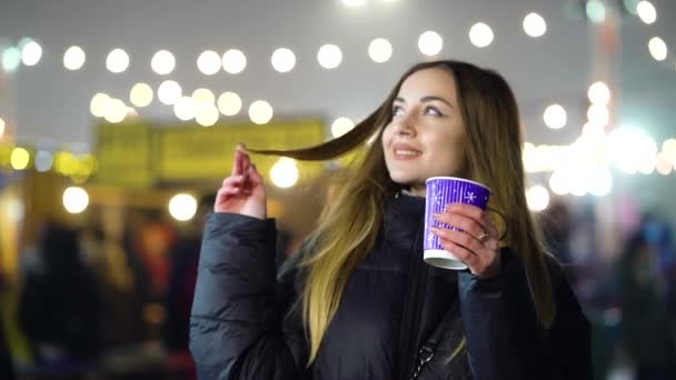 Chica romántica calentando con bebida caliente cerca de pista de patinaje en la noche — Vídeos de Stock