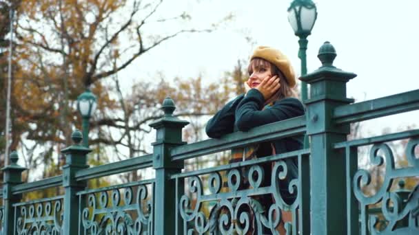 Позитивная девушка в пальто и берете стоит на мосту в осеннем парке — стоковое видео