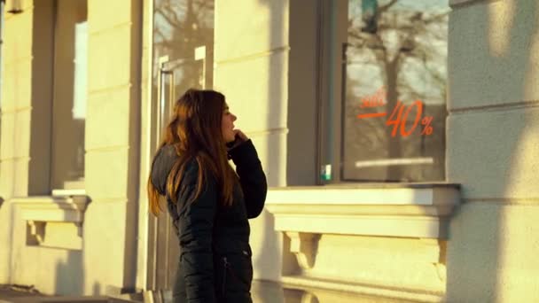 Девушка разговаривает по телефону и смотрит на витрину магазина с табличкой продажи — стоковое видео