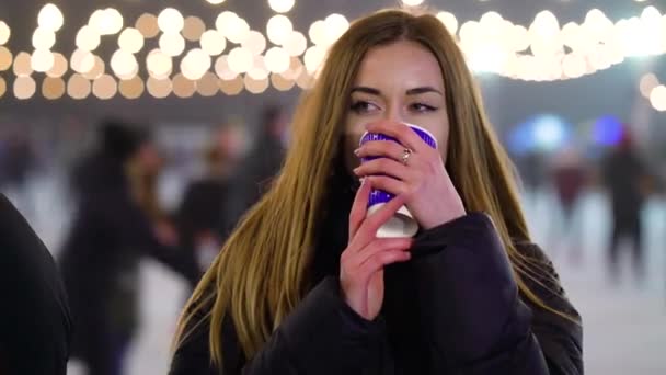 Dreamy girl warming with hot drink near illuminated skating rink in winter — Stock Video