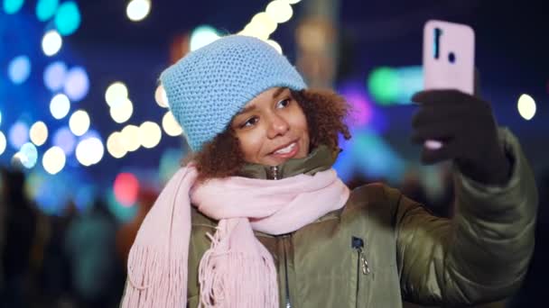 Счастливая чернокожая девушка записывает видео на рождественском базаре ночью — стоковое видео