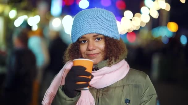 Mooi zwart meisje met warm drankje op de kerstmarkt 's nachts — Stockvideo