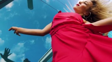 Low angle of free style dance by blonde woman in red dress against glass wall