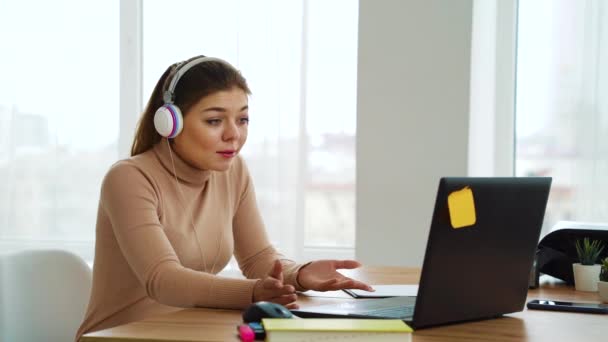 Веселая девушка в наушниках проводит видеоконференцию на ноутбуке в офисе — стоковое видео