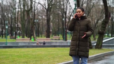 Positive black girl talking on phone in park in dull weather