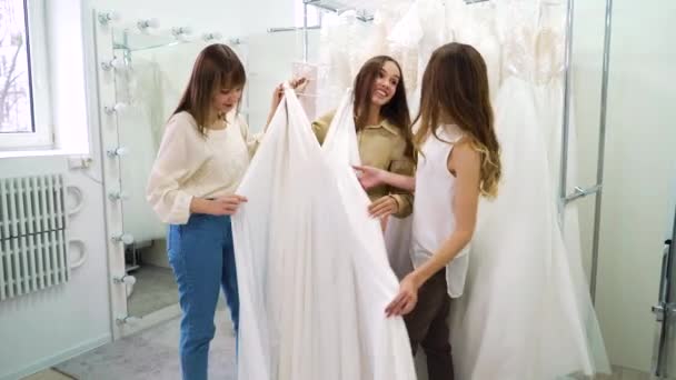 Sposa eccitata con damigelle d'onore cercando di elegante vestito bianco nel salone di nozze — Video Stock