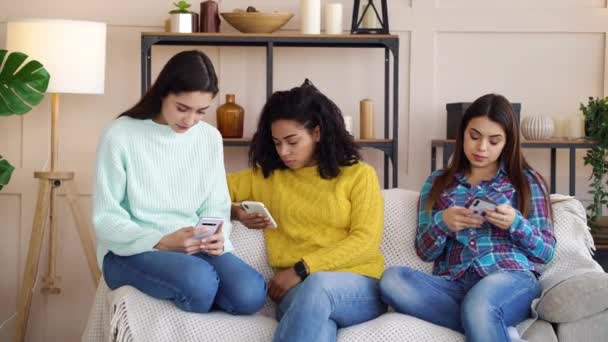 Багаторасові дівчата сидять у вітальні і використовують смартфони — стокове відео