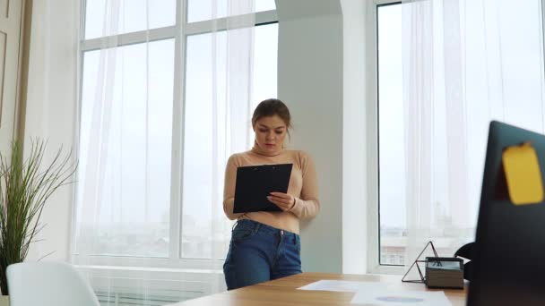 Mujer de negocios joven de pie cerca de la ventana y la escritura en el portapapeles — Vídeos de Stock