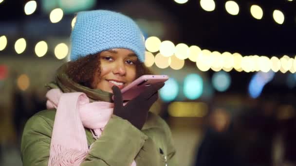 Chica alegre negro hablando por teléfono cerca de pista de patinaje iluminado por la noche — Vídeo de stock