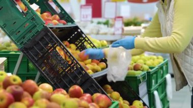 Tıbbi eldivenli bir müşteri marketteki plastik kutulardan elma topluyor.